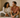 A loving couple sitting on a cozy bed, smiling and laughing together in soft morning light. Warm tones, cozy bedroom, minimalist style, intimate but wholesome atmosphere