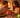 A couple sits on a bed surrounded by candles and rose petals, exchanging a wrapped gift and smiling affectionately.